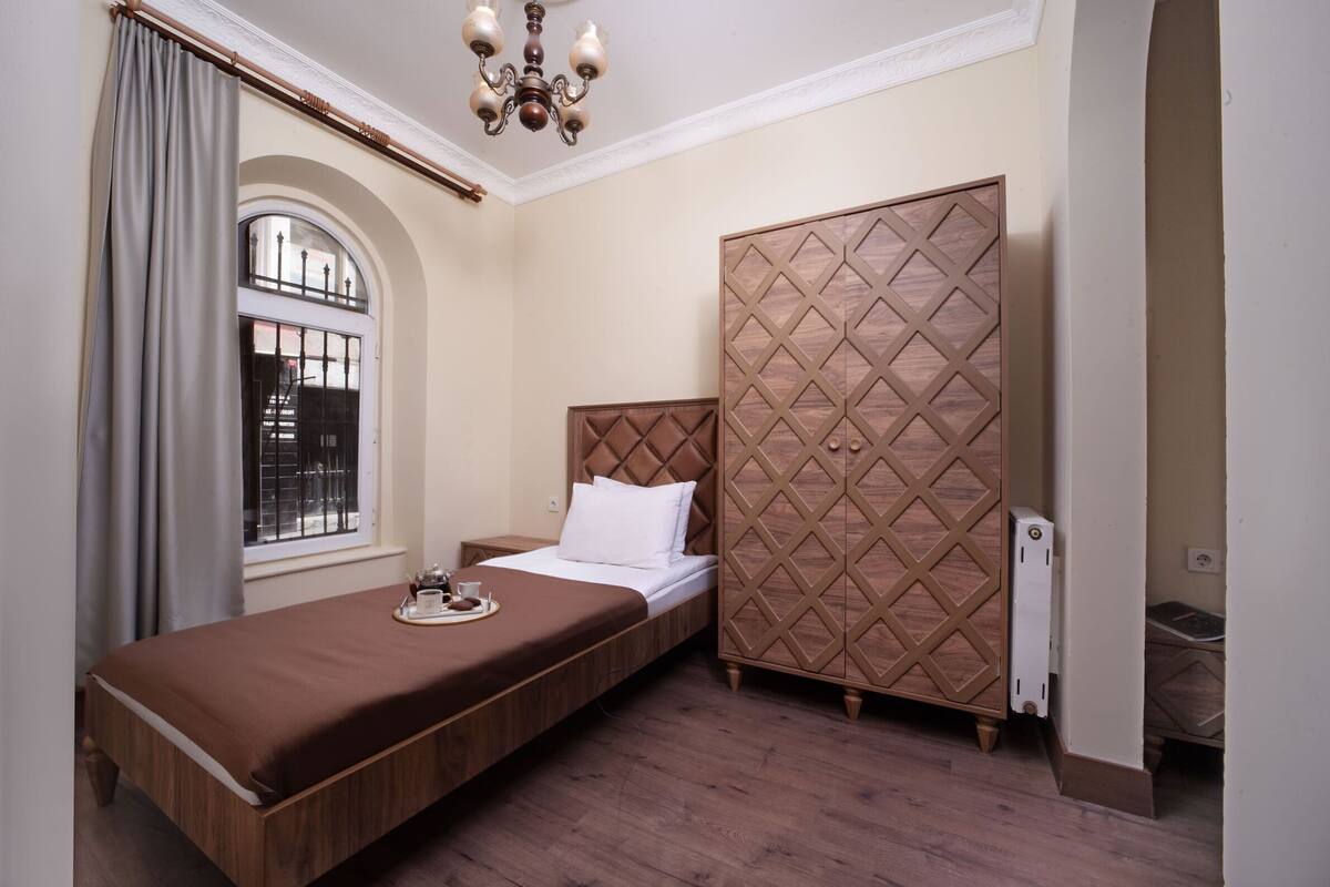 A single bed with a wooden headboard, a bedside table with a tray, a window with curtains, and a wardrobe with a geometric pattern.
