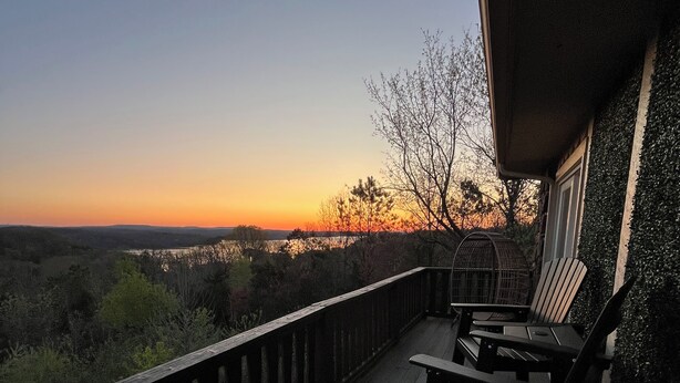 Terraza de madera con barandilla, dos sillas Adirondack y vistas a un paisaje al atardecer.