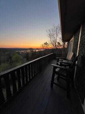 A wooden deck with a railing, two Adirondack chairs, and a view of a sunset over a landscape.