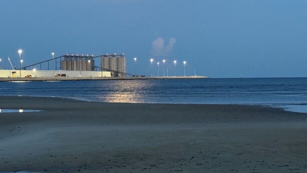 A beach with a pier extending into the water, featuring industrial structures and lights.