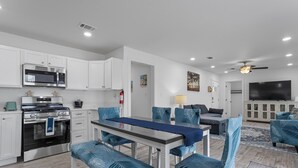 A modern kitchen with white cabinets, a stainless steel stove, and a microwave. There is a dining area with blue upholstered chairs and a table.
