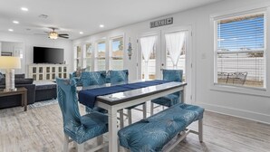 A spacious dining area with a white table and blue upholstered chairs, a ceiling fan, and a TV mounted on the wall.