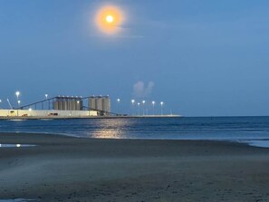 Une plage avec une grande structure industrielle en arrière-plan, une étendue d’eau et un ciel dégagé.