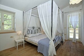 Una cama con dosel y cuatro columnas, una mesita de noche con lámpara, y un ventanal con cortinas.