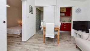 Un espacio habitable compacto con cama, una mesa de comedor con sillas, una zona de cocina con armarios rojos, un televisor y un reloj en la pared.