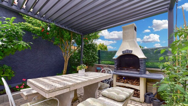 Un patio exterior con una mesa de piedra, sillas acolchadas, una parrilla empotrada y vistas al campo.