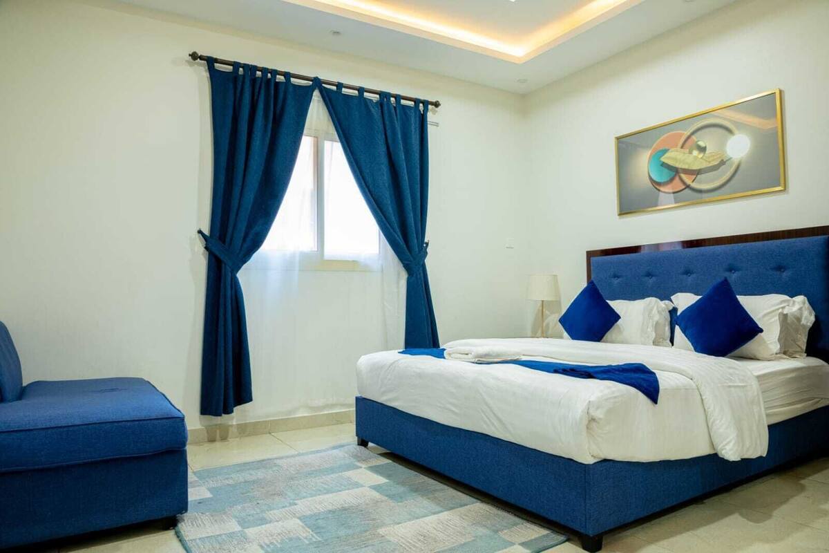 A hotel room with a bed, a blue armchair, and a painting on the wall.