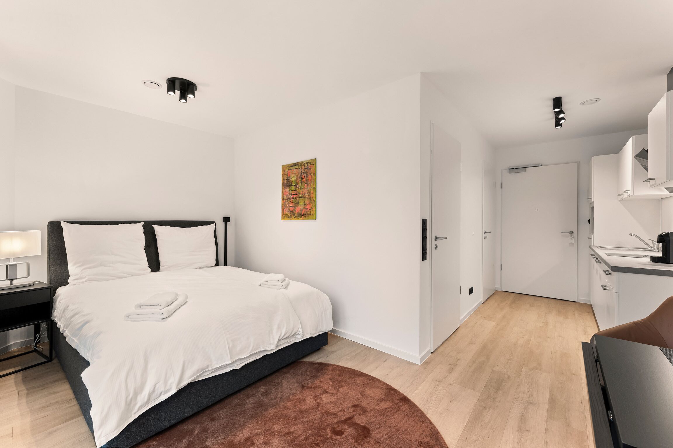 A modern bedroom with a bed, a nightstand, a desk, and a door leading to a bathroom.