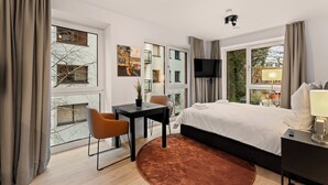 Habitación de hotel moderna con una cama grande, un escritorio con sillas, un televisor y vistas a la ciudad a través de amplias ventanas.