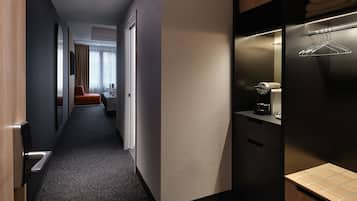 Pasillo de hotel con una cama, un área de descanso y una kitchenette con cafetera.