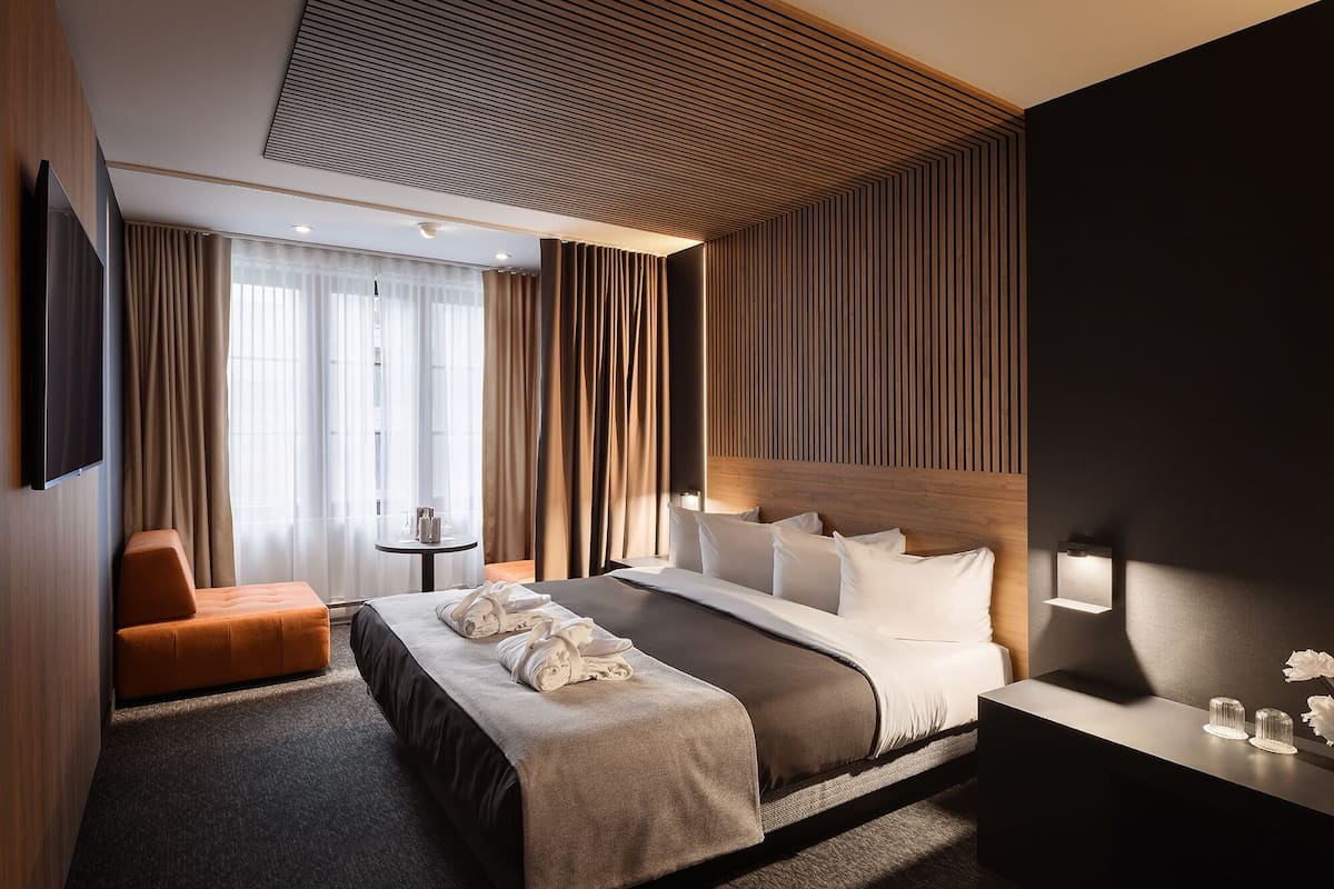 A modern hotel room with a large bed, a bedside table, a small table with a lamp, and a seating area with a chair.