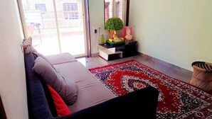 A living room with a sofa, a red patterned rug, a small table with a Buddha statue, and a wicker chair. - Shri Swami Samarth Home Stay (Akalkot)