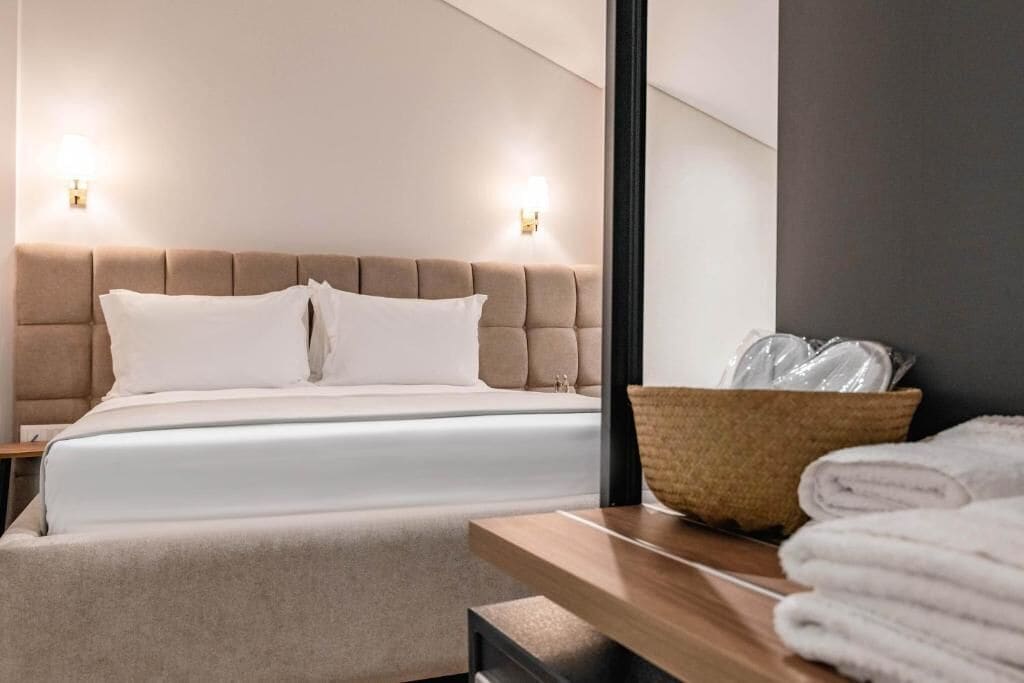 Quarto de hotel moderno com uma cama grande, duas luminárias de cabeceira, uma mesinha de madeira e um cesto de palha com toalhas.