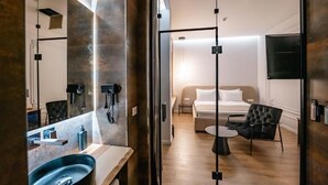 Ein modernes Hotelzimmer mit einem großen Spiegel, einem Waschbecken, einem Bett, einem Sessel und einem Fernseher.