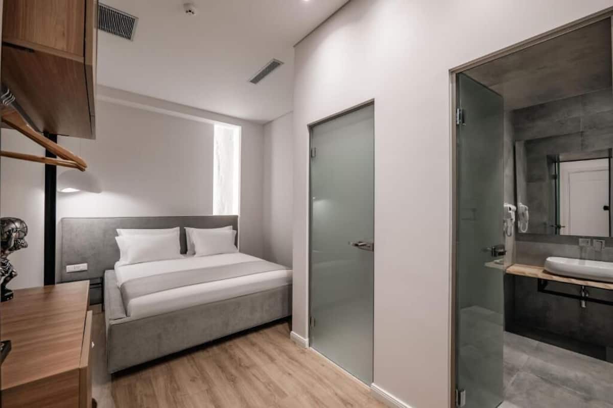 A modern bedroom with a bed, a bedside table, a wardrobe, and a glass-enclosed bathroom with a sink and mirror.