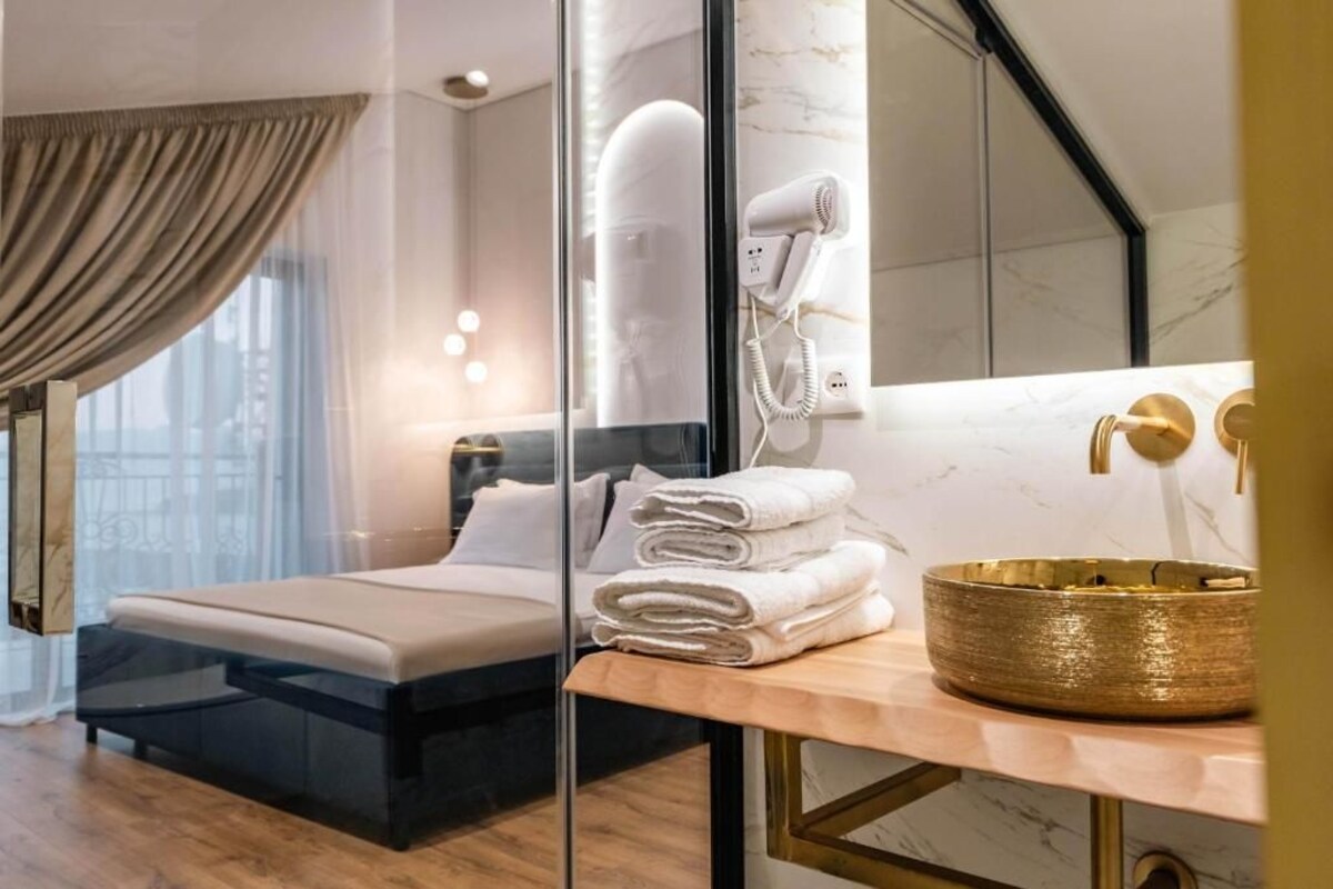 Et moderne hotelværelse med en seng, en glasvæg, et toiletbord med vask i guld og en hårtørrer på væggen.