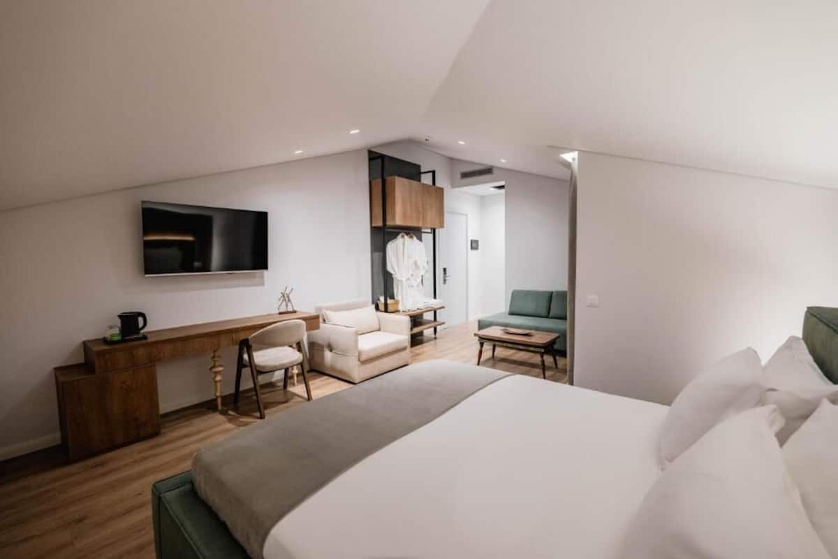 A modern hotel room with a large bed, a desk, a chair, a sofa, and a TV mounted on the wall.