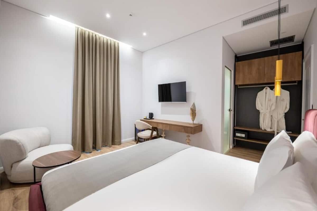 A modern hotel room with a large bed, a desk, a chair, a TV, and a wardrobe.
