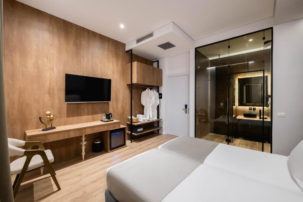 A modern hotel room with a wooden accent wall, a flat-screen TV, a desk with a chair, a bed, and a walk-in closet with a mirror.
