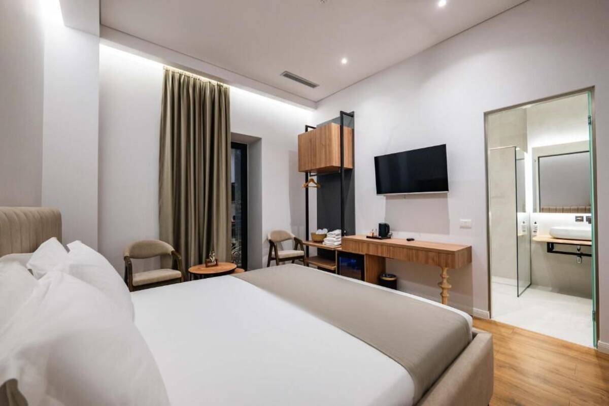 A modern hotel room with a large bed, a desk, a chair, a TV, and a bathroom visible through a glass door.