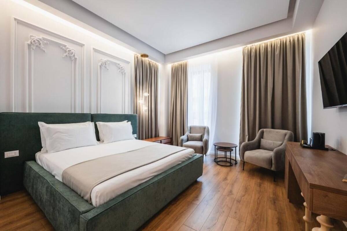 A modern hotel room with a large bed, two armchairs, a small round table, and a TV on the wall.