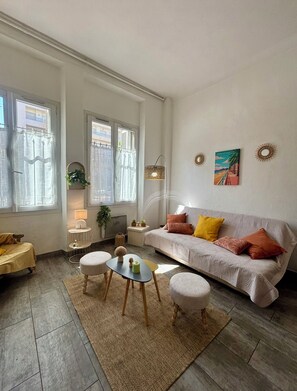 Un salon avec un canapé, une table basse et un tapis. Il y a des fenêtres avec des rideaux, une lampe et un tableau au mur.