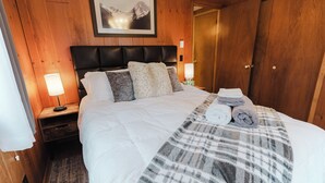 Un lit bien fait, avec une couverture à carreaux, des oreillers et des serviettes enroulées. On y voit une tête de lit en bois, une table de chevet avec une lampe et un tableau encadré accroché au mur.