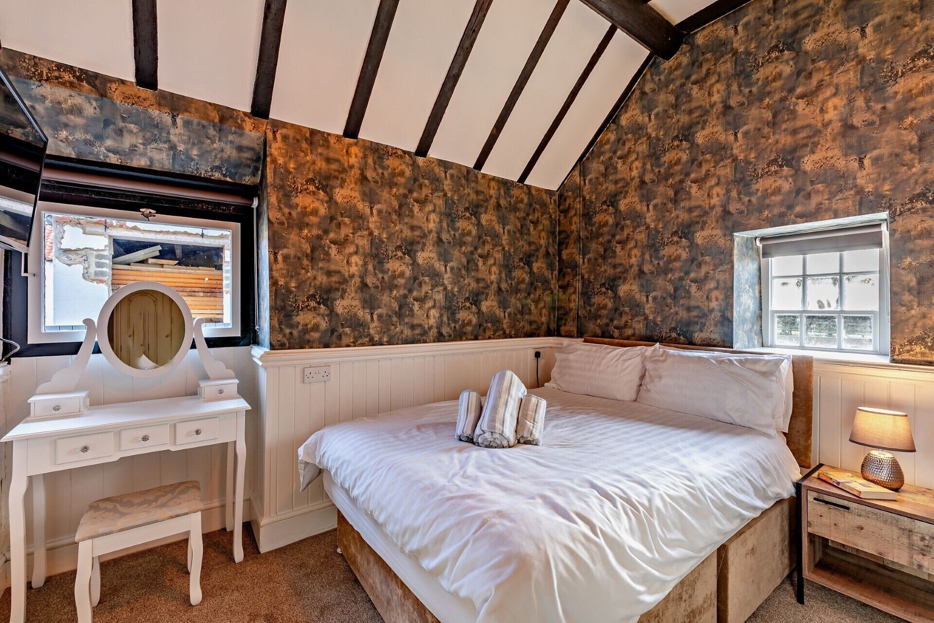 A bedroom with a large bed, a vanity with a mirror, a window, and a lamp on a bedside table.