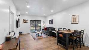A modern living room with a sofa, coffee table, and dining table. There is a TV mounted on the wall and a large window with a view of the outdoors. - Gateway to Alaska Wonders A308 (Juneau)