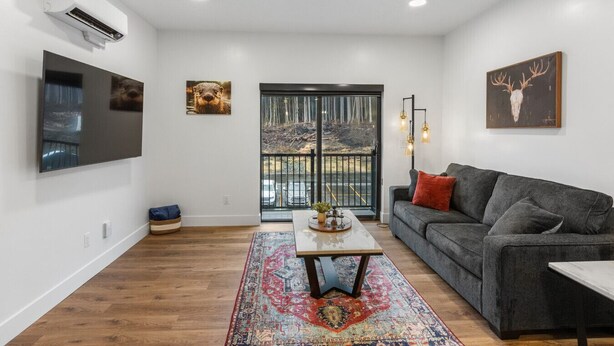 A modern living room with a grey sofa, a coffee table, and a large TV mounted on the wall. There is a large window with a view of trees and a parking lot. - Gateway to Alaska Wonders A308 (Juneau)