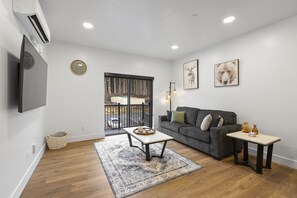 Una sala de estar moderna con un sofá gris, una mesita de centro, una mesita auxiliar y un ventanal con vista a un estacionamiento.