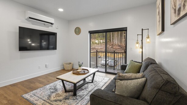 A living room with a flat-screen TV, a sofa, a coffee table, and a large window with a view of trees. - Juneau Jewel: Luxury & Relaxation A307 (Juneau)