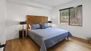A bedroom with a wooden headboard, a bed with a blue bedspread, two bedside lamps, and a window with a forest view. - Juneau Jewel: Luxury & Relaxation A307 (Juneau)