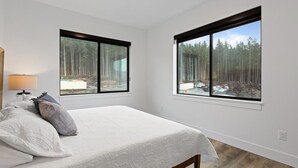 A bedroom with a bed, two windows, a lamp, and a view of a forest. - Affordable Luxury With Views!  A305 (Juneau)