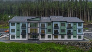 Un edificio de varios pisos con una entrada central, balcones y un fondo boscoso.