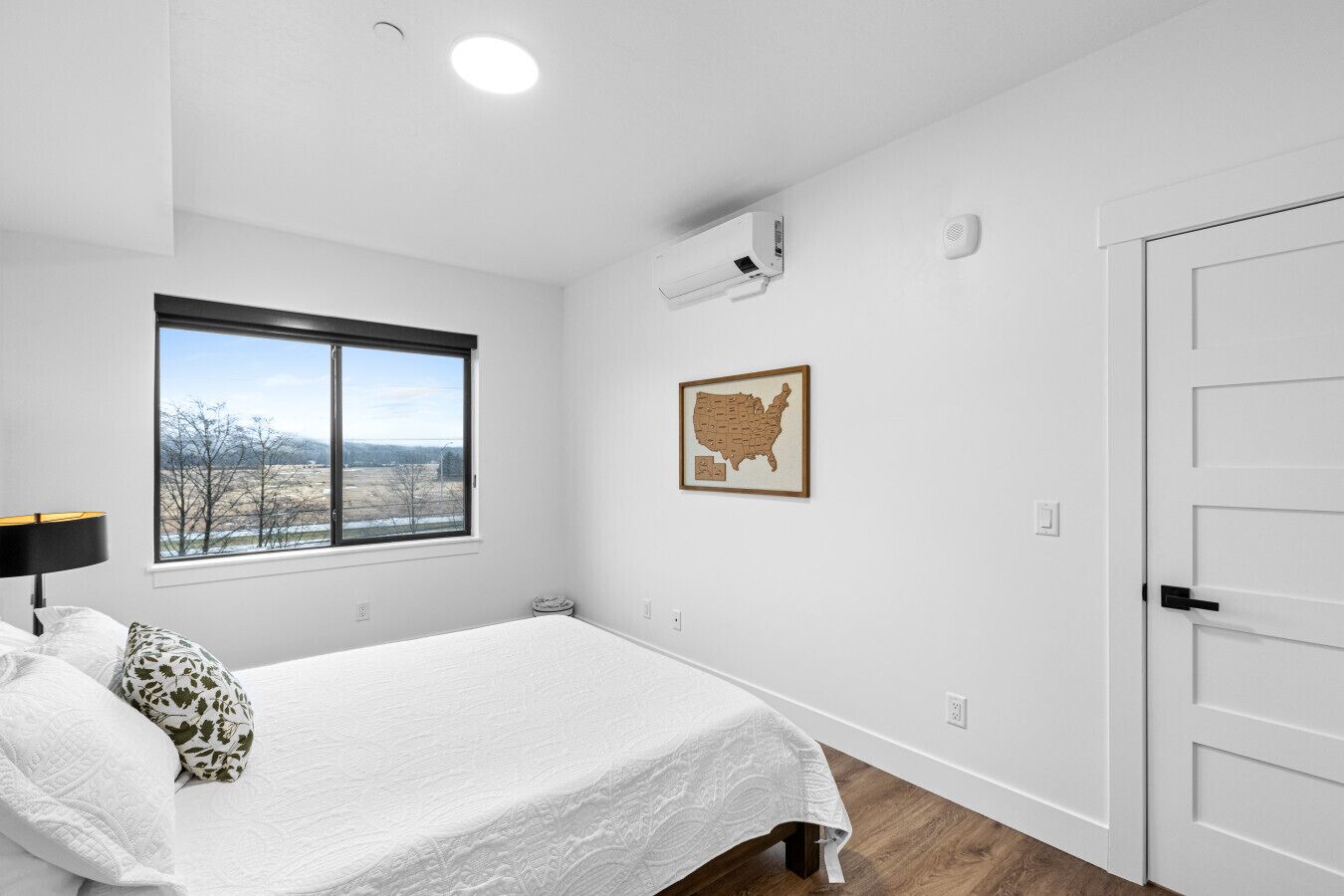 A bedroom with a bed, a window with a view, a wall-mounted air conditioner, a framed map, and a door.