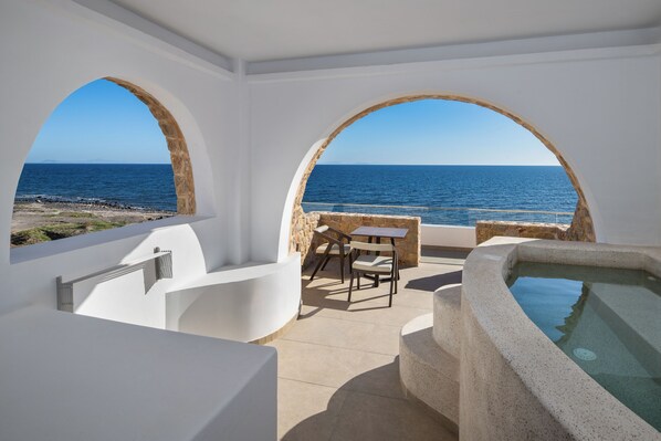 Une chambre moderne dotée d’une grande fenêtre en arc de cercle donnant sur la mer, d’une baignoire et d’une petite table avec des chaises.