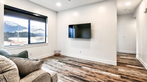 A modern living room with a large window, a flat-screen TV mounted on the wall, and a wooden floor.