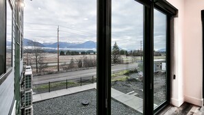A view from a window overlooking a road with mountains in the distance, a clear sky, and a building with a green facade. - Gorgeous Views, Cozy Luxury 209 (Juneau)