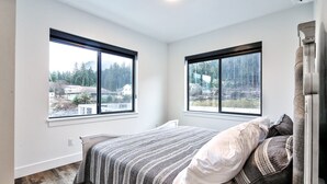 A bedroom with a bed, two windows, and a view of a forested area. - Gorgeous Views, Cozy Luxury 209 (Juneau)