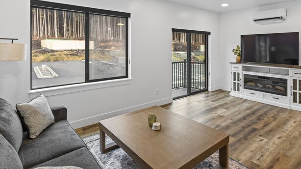 A modern living room with a sofa, coffee table, TV, and large windows overlooking a yard. - Cozy, Spacious Retreat with Fireplace 205 (Juneau)