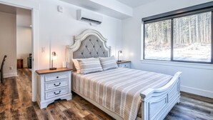 A bedroom with a bed, nightstands, an air conditioner, a window with a view of trees, and a door leading to another room. - Cozy, Spacious Retreat with Fireplace 205 (Juneau)