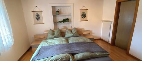 Un dormitorio con techo de madera, cama con ropa de cama verde y gris, una pequeña estantería con plantas y un radiador.