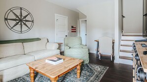 Una sala de estar con una mesa de centro de madera, un sofá, un sillón verde claro, una alfombra, una brújula montada en la pared y una escalera que lleva a otro nivel.
