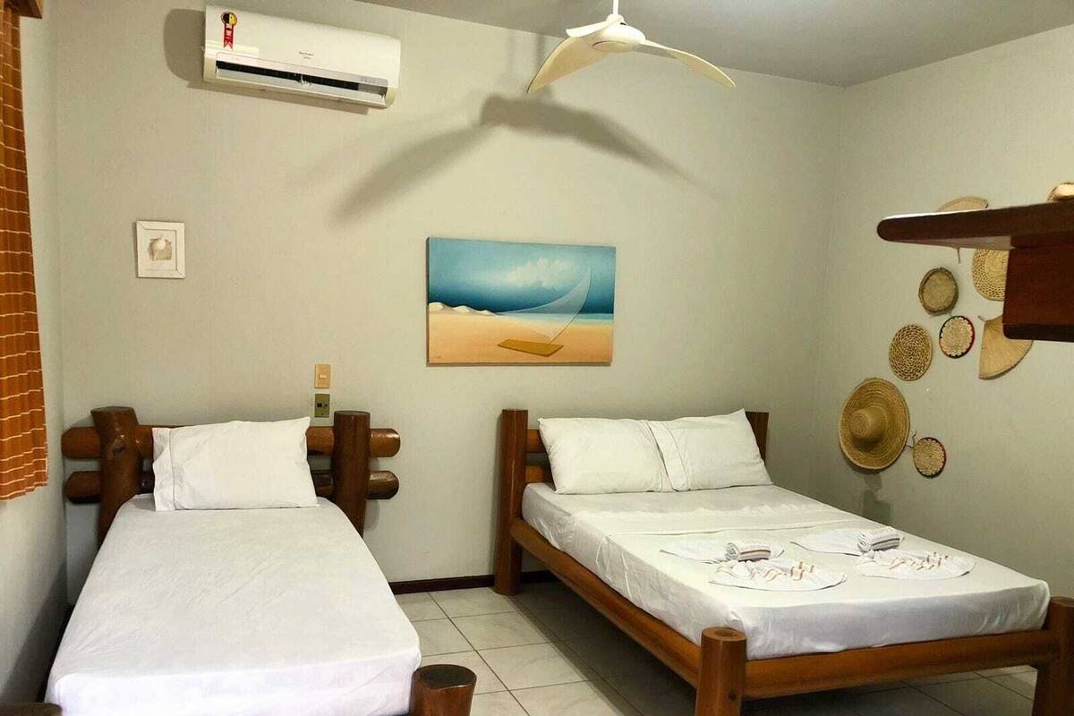 Una habitación con dos camas de madera, un aire acondicionado, un ventilador de techo, un cuadro de playa y una pared decorada con sombreros tejidos.