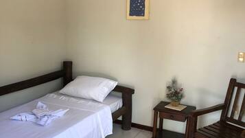 Ein Einzelbett mit Holz-Kopfstück, ein kleiner Tisch mit Pflanze und ein Holzstuhl in einem Raum mit Fliesenboden und Deckenventilator.