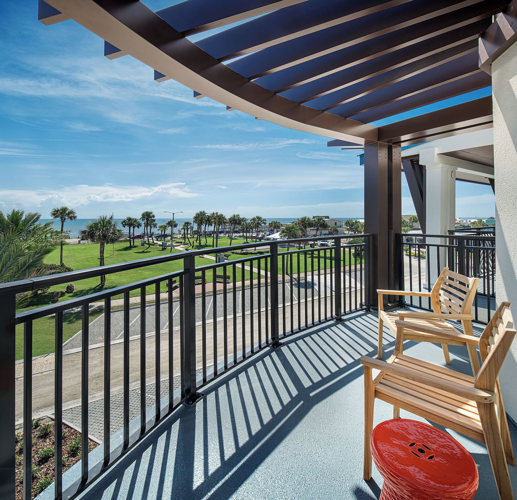 A balcony with a view of a golf course, a red stool, and two wooden chairs.