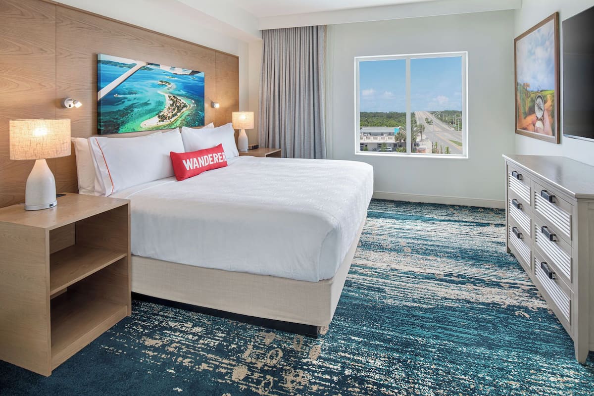 A hotel room with a large bed, a nightstand, a dresser, a painting of a tropical scene, a window with a view of a road and greenery, and a lamp.