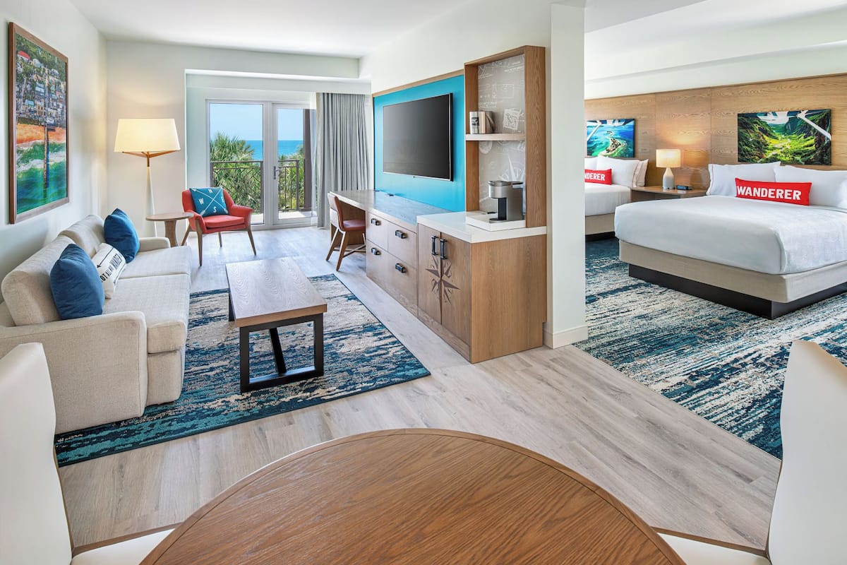 A hotel room with a bed, a desk, a sofa, a dining table, and a view of the ocean.