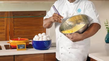 Un chef con uniforme blanco batiendo huevos en un bol. Hay un bol azul con huevos y un recipiente amarillo etiquetado como 'Scrambled Eggs' sobre la encimera.